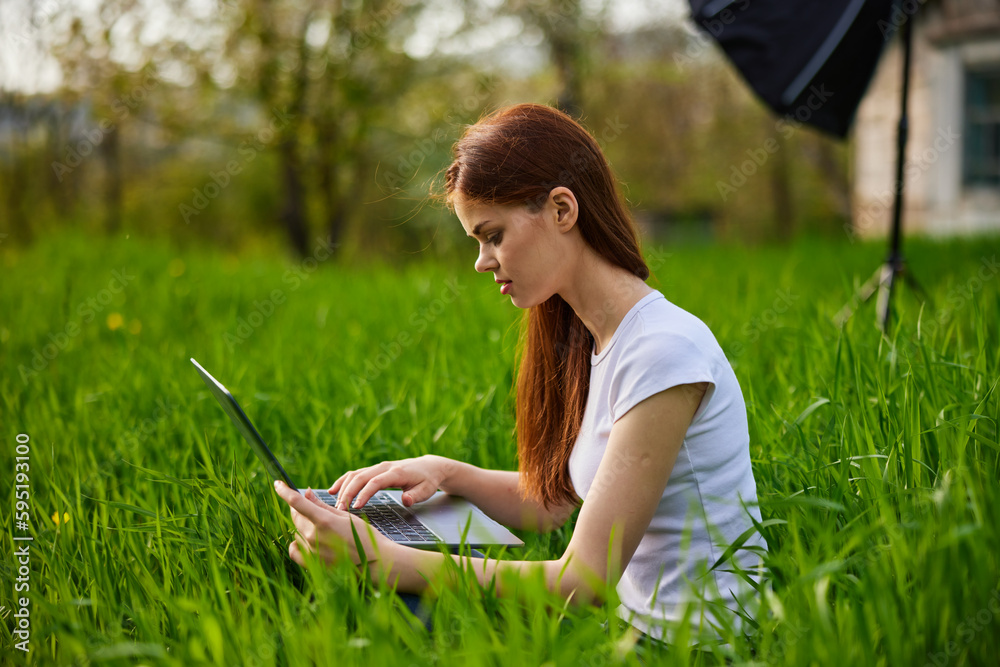 a woman in a light T-shirt sits in high green grass working at a laptop