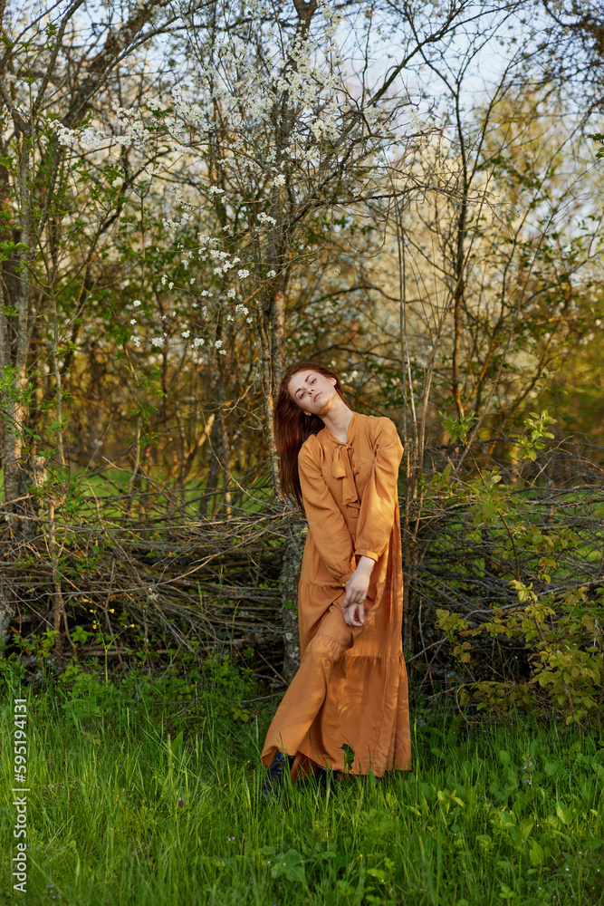 an elegant, sophisticated woman poses relaxed standing near a wicker fence at the dacha in a long orange dress, looking pleasantly at the camera an elegant, sophisticated woman poses relaxed standing near a wicker fence at the dacha in a long orange dress, looking pleasantly at the camera