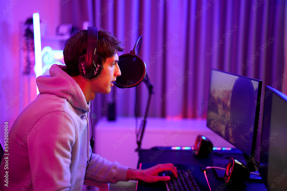 Caucasian attractive gamer boy typing keyboard while playing shooting game online with his team in violet neon light room at the apartment Caucasian attractive gamer boy typing keyboard while playing shooting game online with his team in violet neon light room at the apartment