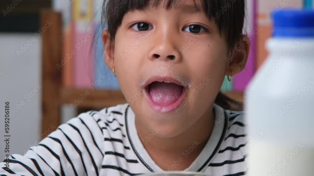 Cute Asian girl drinking a glass of milk at home in living room. Little girl drinking milk in the morning before going to school. Healthy food in childhood. Cute Asian girl drinking a glass of milk at home in living room. Little girl drinking milk in the morning before going to school. Healthy food in childhood.