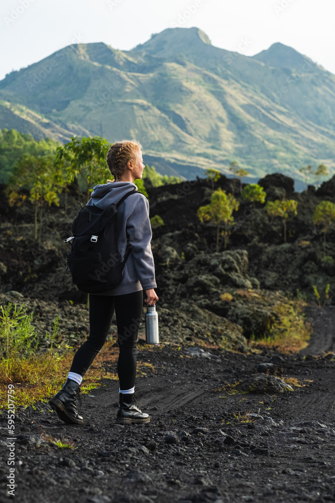 A young girl traveler with a backpack walks in the mountains against the backdrop of the Batur volcano on the island of Bali in Indonesia.