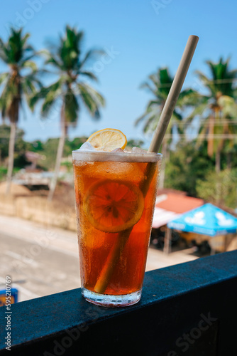 A tall glass with a refreshing  cold iced tea. Orange-brown cocktail ice tea with ice cubes and a slice of lemon as decoration.