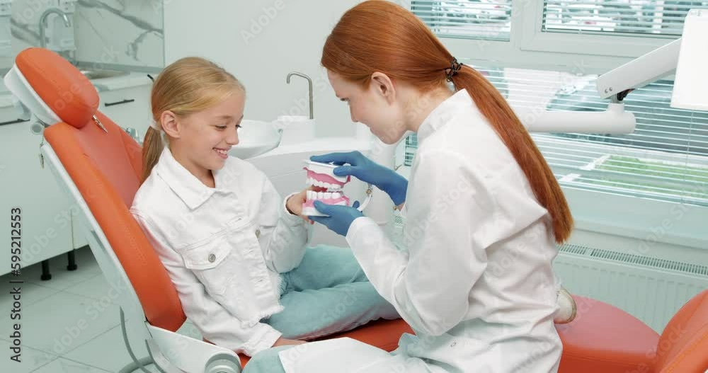 friendly kind smiling stomatologist in uniform with maket of jaw is playing with blonde little girl , reliving stress, creating positive mood , telling about healthcare game entertainment lifestyle friendly kind smiling stomatologist in uniform with maket of jaw is playing with blonde little girl , reliving stress, creating positive mood , telling about healthcare game entertainment lifestyle