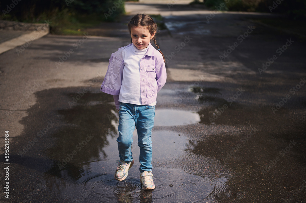 Full length portrait of a beautiful mischievous happy little kid girl in jeans and purple jacket, smiling and having fun, jumping on a small puddle after rain while walking on the countryside street.
