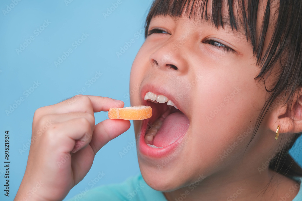 Smiling cute little girl eating sweet gelatin with sugar added isolated on blue background. Children eat sugary sweets, causing loss teeth or tooth decay and unhealthy oral care. Smiling cute little girl eating sweet gelatin with sugar added isolated on blue background. Children eat sugary sweets, causing loss teeth or tooth decay and unhealthy oral care.