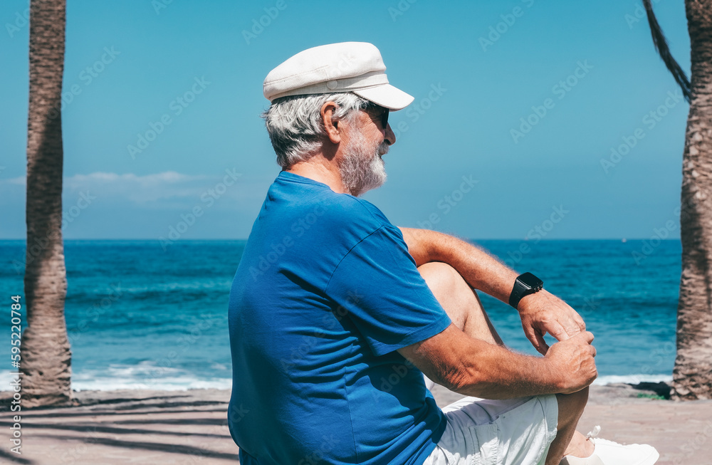 Side View of Smiling Senior Man Sitting at the Beach Looking Away. Portrait of Retiree Caucasian Man Relaxing Enjoying Sunny Day at Sea