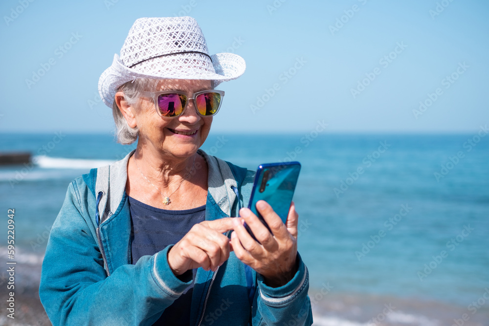 Smiling old senior woman in hat and sunglasses sitting outdoors at sea typing message on mobile phone, elderly lady enjoying sunny day, vacation or retirement concept. Horizon over water