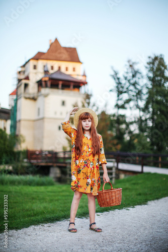 a little girl with long red hair in a yellow dress is standing in front of the old Castle Blatna in Czech Republic and holds a basket in a hand