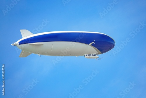 A blue and white airship with passengers on board flies against the blue sky. 