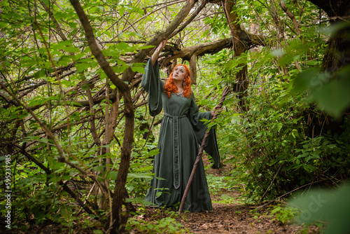 A young woman in a cloak, with a staff and a green dress in the form of a druid. Forest witch with bright red hair. Makeup with black pencil.