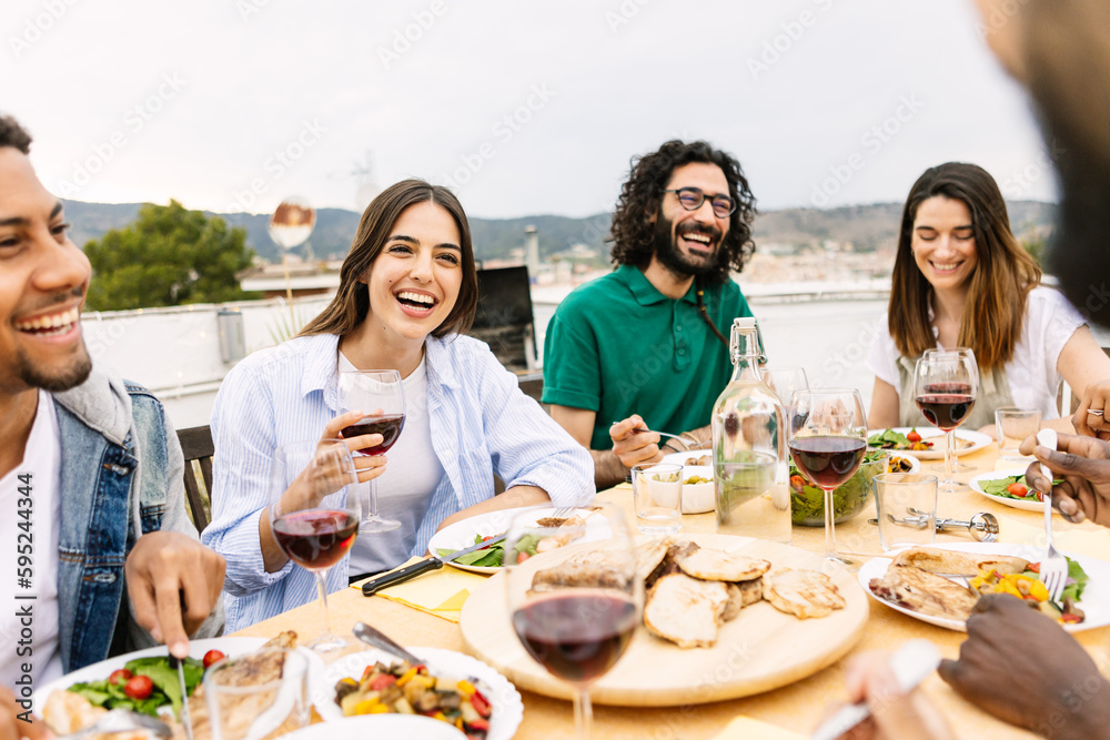 Smiling group of diverse friends having dinner at rooftop party. Happy millennial multiracial people laughing and having fun while drinking and eating sitting on table in summer celebration. Smiling group of diverse friends having dinner at rooftop party. Happy millennial multiracial people laughing and having fun while drinking and eating sitting on table in summer celebration.