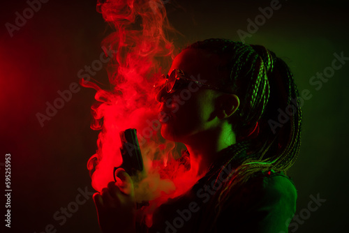 A young woman with pigtails in the dark in a club in round dark glasses holds a gun in her hands. Vaping in the city. Girl with dreadlocks in smoke. red green color