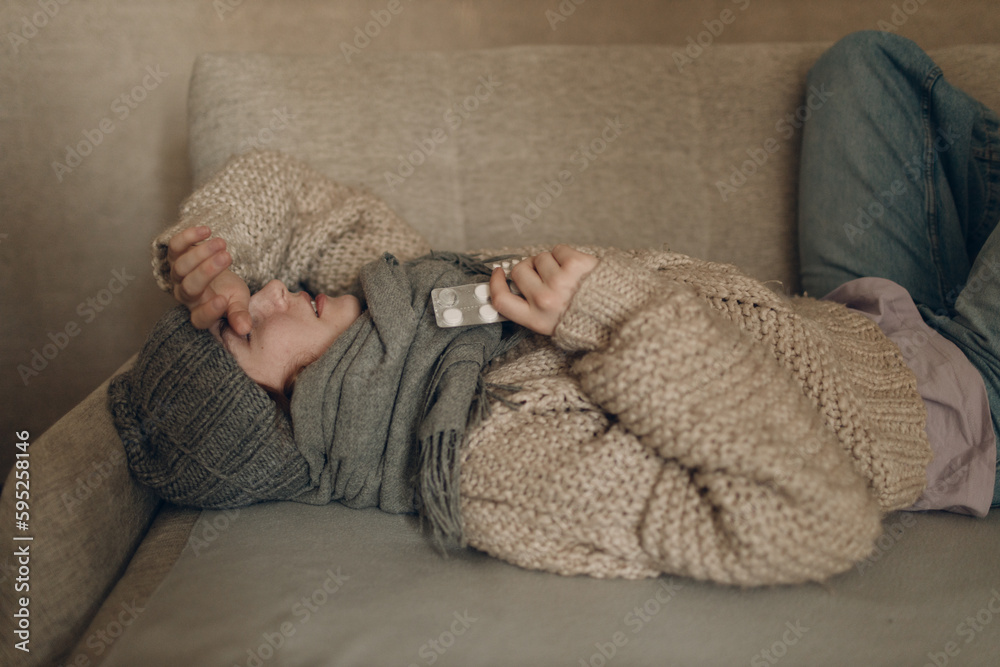 Young woman wearing scarf holding pills in hands has flu ill sick disease cold at home indoor on sofa Young woman wearing scarf holding pills in hands has flu ill sick disease cold at home indoor on sofa