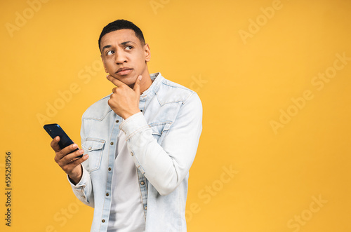 Serious thinking african american young man holding phone isolated on yellow studio background with copy space aside, african guy using mobile applications, texting. © denis_vermenko