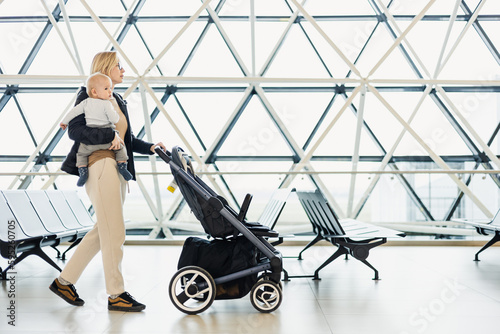 Mother carying his infant baby boy child, pushing stroller at airport departure terminal moving to boarding gates to board an airplane. Family travel with baby concept