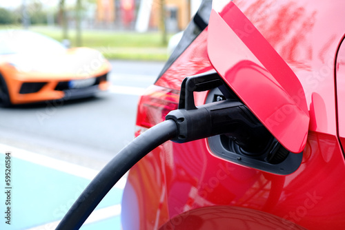 car in red color on street, charging with electricity through cable, electric vehicle in European city, energy cylindrical accumulators, alternative energy development, technology and innovation