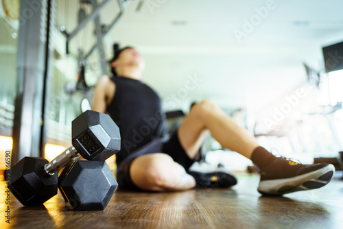 Happy cheerful Asian sportsman relaxing - sitting on the floor after done a weight training exercise in an indoor fitness or gym. Man resting after did a wight training workout.