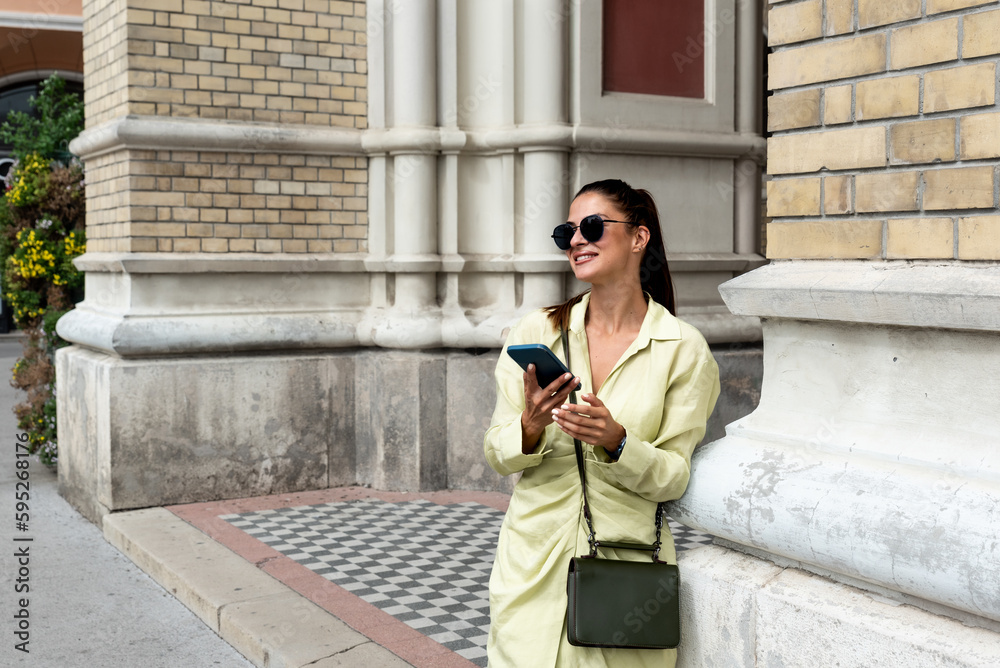Fototapeta premium Young beautiful smiling stylish woman. Sexy carefree model posing in the street. Positive brunette female holding smartphone. Looking at cellphone screen. Using phone apps. Outdoors at sunny day