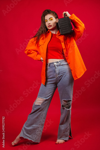A young girl in a bright orange shirt and jeans on a red background holds a tape recorder from the 90s in her hands.