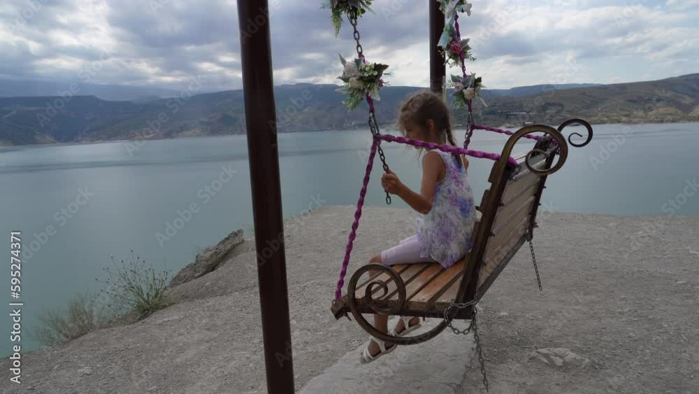 Cute little girl in dress with her hands raised in delight, riding on swing decorated with flowers and sequins on cliff of river bank in summer Cute little girl in dress with her hands raised in delight, riding on swing decorated with flowers and sequins on cliff of river bank in summer