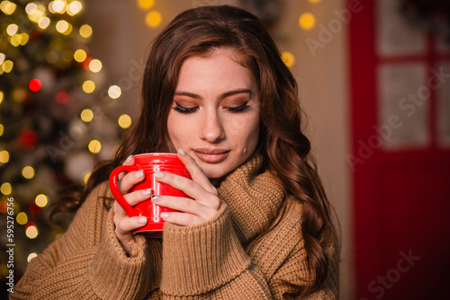 A young woman in a beige sweater sits in a Christmas decoration against the backdrop of a Christmas tree and lights. The girl is holding a red mug in her hands.