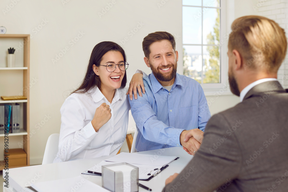 Fototapeta premium Successful deal. Husband and wife receive congratulations from bank employee on renting, buying or mortgage. Happy woman makes joyful fist while her husband shakes hands with financial advisor.
