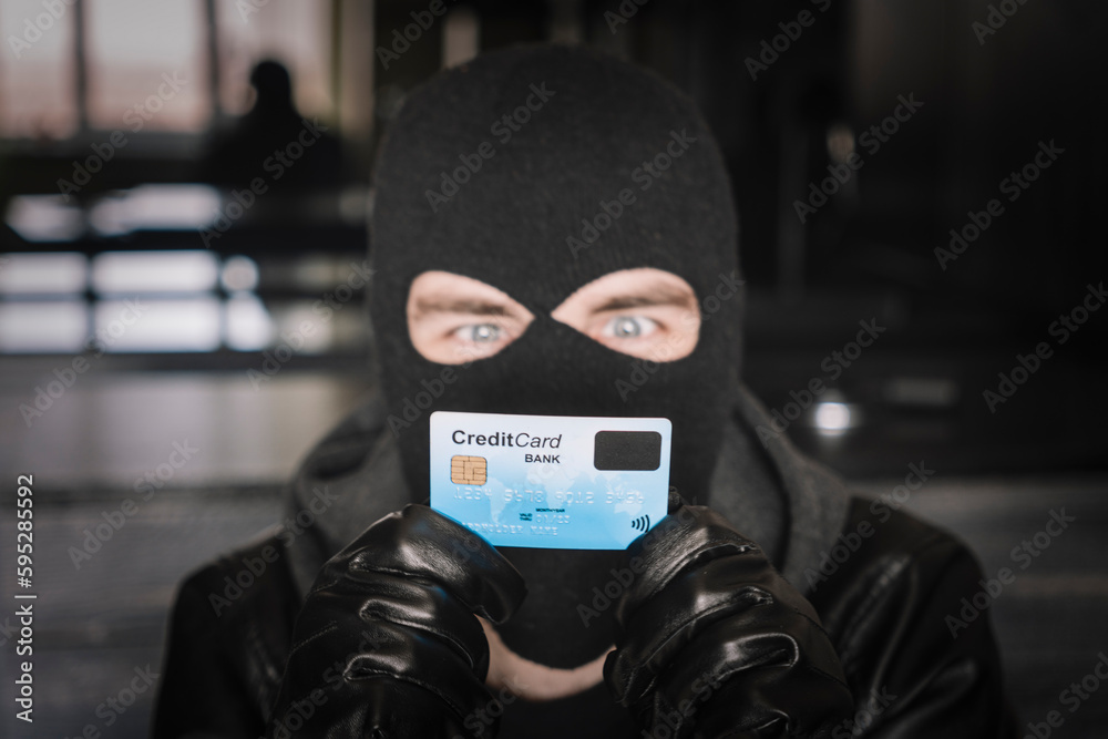 A thief in a black mask holds a stolen bank card in his hands. Skilled masked criminal using a stolen credit card to buy things online. A thief in a black mask holds a stolen bank card in his hands. Skilled masked criminal using a stolen credit card to buy things online.