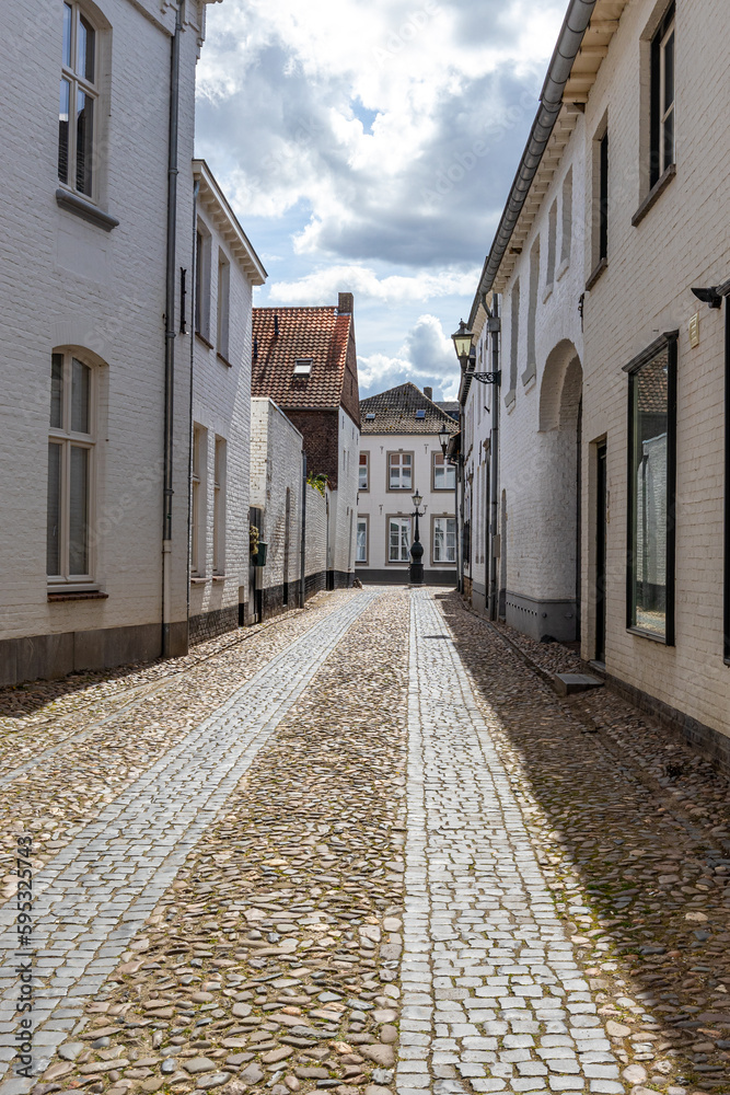 Landscape of narrow cobbled street between house with white walls lantern and building in background against blue sky and clouds, gabled roofs, old Dutch town of Thorn in Midden-Limburg, Netherlands