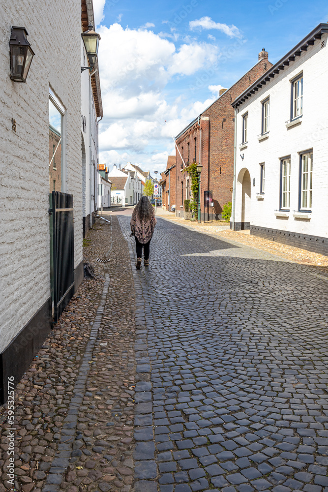 Cobbled street between houses with white and brick walls against blue sky and clouds, adult woman walking with her back to camera, sunny day in old Dutch town of Thorn in Midden-Limburg, Netherlands
