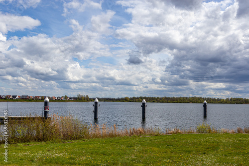 Metal poles next to pier and reinforcement of Thorn Wessem dike on Maas river  cityscape and trees in background against blue sky with white clouds  sunny spring day in Midden-Limburg  Netherlands