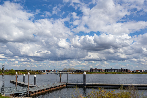 Pier for boarding tourist boats and Thorn Wessem dyke reinforcement on river Maas, townscape in background against blue sky covered with white clouds, sunny spring day in Midden-Limburg, Netherlands © Emile