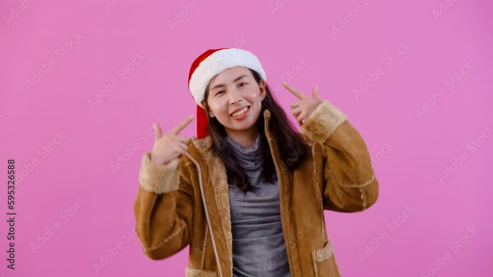 4K, Beautiful Asian girl in long-sleeved shirt wearing Christmas hat, swaying back and forth, both hands pointing to his own face, happy expression, Isolated indoor studio on pink background. 4K, Beautiful Asian girl in long-sleeved shirt wearing Christmas hat, swaying back and forth, both hands pointing to his own face, happy expression, Isolated indoor studio on pink background.