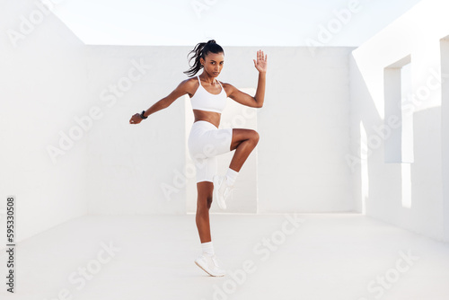 Young woman athlete jumping in white outdoor studio. Slim female doing cardio exercises. Sportswoman practicing high-intensity exercises.