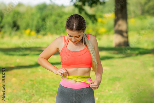 Active young excited woman with centimeter measuring tape on her slim waist in park. Sport, fitness, healthy lifestyle and diet concept.