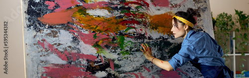 Creative young female painter working on a large modern abstract oil painting, applying paint on canvas with fingers in living room at home studio workshop