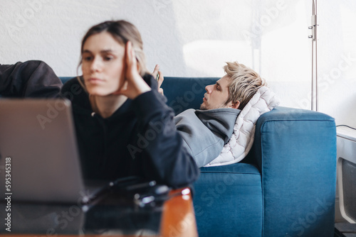 Woman work on laptop, man lies on blue sofa and read news in mobile phone. Couple spend time separately in they house. Resting free time. Focus on man.