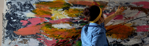 Back view of young woman, female painter in apron looking focused while creating a large modern abstract oil painting, applying paint on canvas with fingers