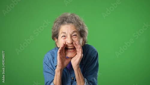 Fotografie Portrait of happy elderly senior old woman with wrinkled skin and grey hair smiling scream and shout calling inviting with hands at mouth say hey you isolated on green screen background