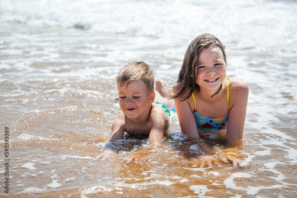 Two children brother and sister (boy and girl) in swimming costumes lying on their stomachs on the sand in the sea water and smiling Two children brother and sister (boy and girl) in swimming costumes lying on their stomachs on the sand in the sea water and smiling