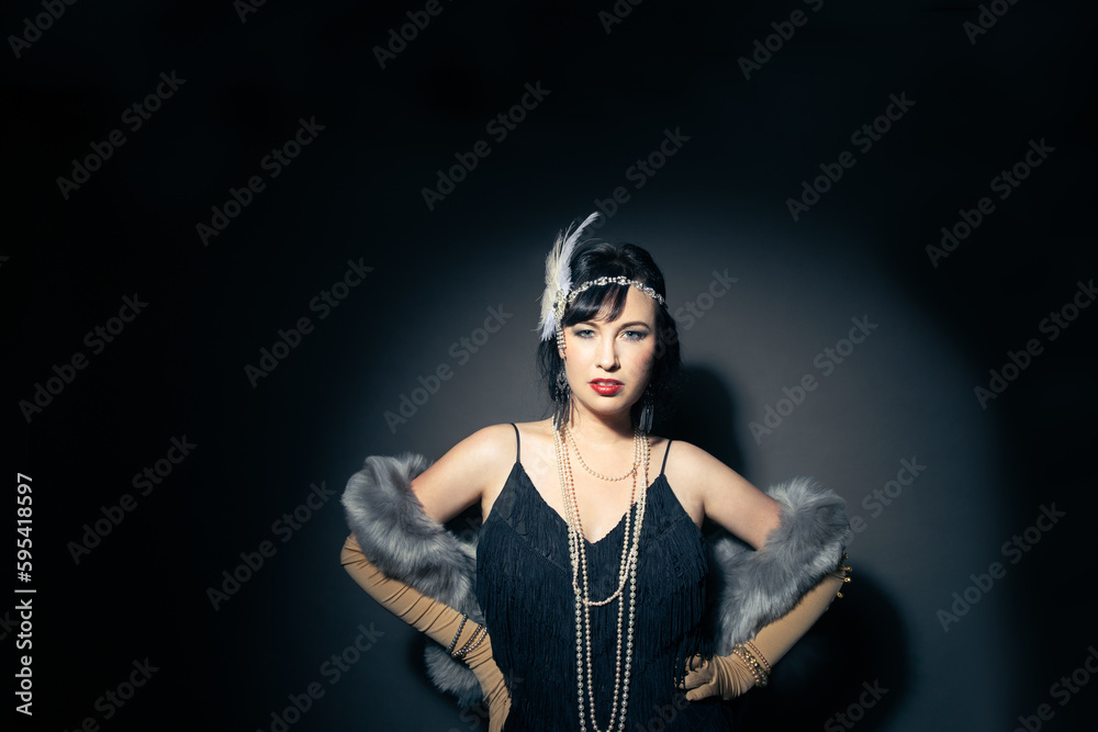 Studio portrait of a beautiful caucasian woman wearing flapper attire. She is standing in a patch of light. She is wearing a fringe trim dress, with elbow length gloves and a blue fur scarf. 