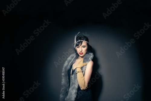 Studio portrait of a beautiful caucasian woman wearing flapper attire. She is standing in a patch of light. She is wearing a fringe trim dress, with elbow length gloves and a blue fur scarf.  © Angela