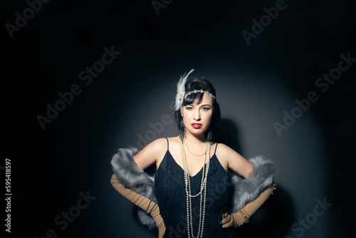 Studio portrait of a beautiful caucasian woman wearing flapper attire. She is standing in a patch of light. She is wearing a fringe trim dress, with elbow length gloves and a blue fur scarf. 