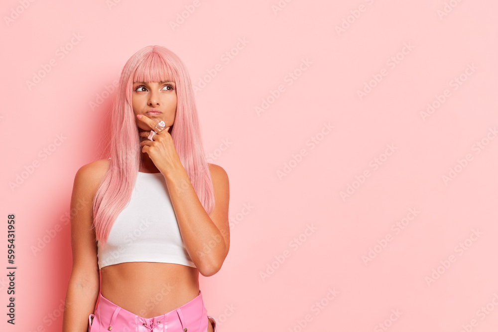 Young woman poses in the middle of pink wall of studio, stands with one hand on her chin, looks up thoughtfully waiting for good news, pause to think concept, copy space, high quality photo Young woman poses in the middle of pink wall of studio, stands with one hand on her chin, looks up thoughtfully waiting for good news, pause to think concept, copy space, high quality photo