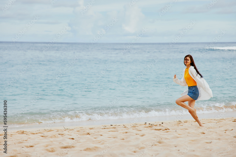 Sports woman runs along the beach in summer clothes on the sand in a yellow T-shirt and denim shorts white shirt flying hair ocean view Sports woman runs along the beach in summer clothes on the sand in a yellow T-shirt and denim shorts white shirt flying hair ocean view