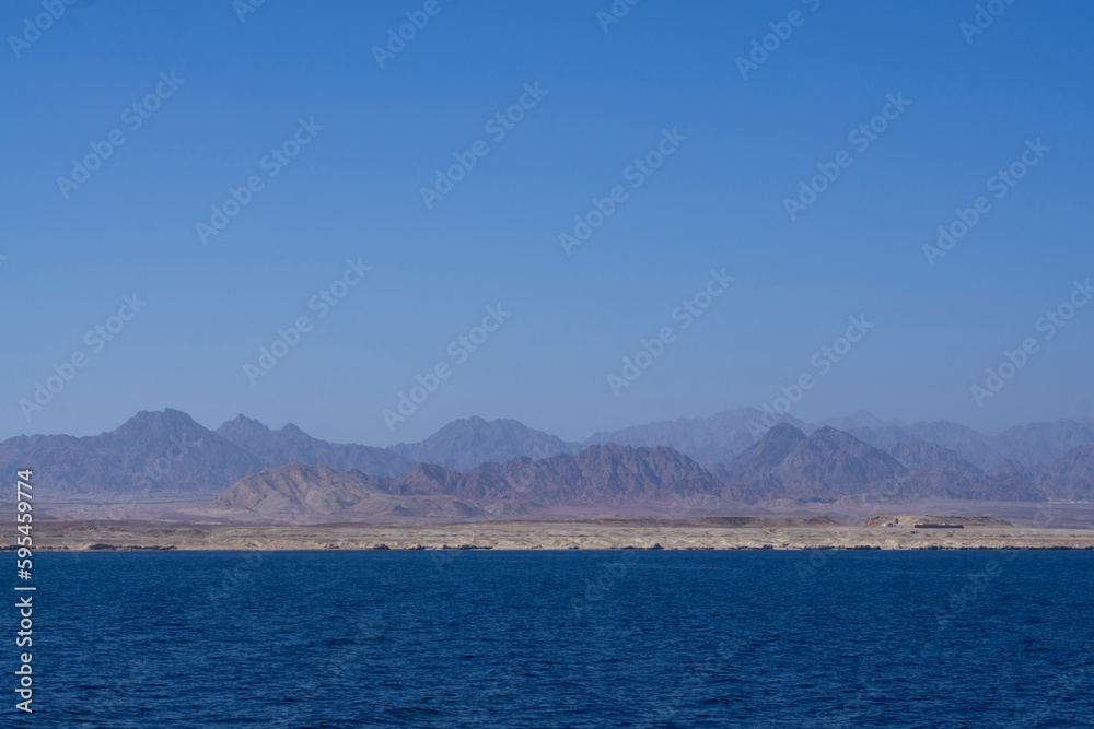 Yachts and boats moored in a sea harbor of Sharm El-Sheikh, view on a coast with panorama of mountains and lighthouse. Mountain landscape with tourist cruise boats near the Ras-Mohammed Reserve.