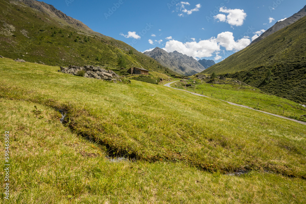 Naklejka premium View of an alpine landscape with a small river in the foreground, and a wooden bard with surrounding mountains in the background during summer. Umhausen, Tyrol, Tirol.