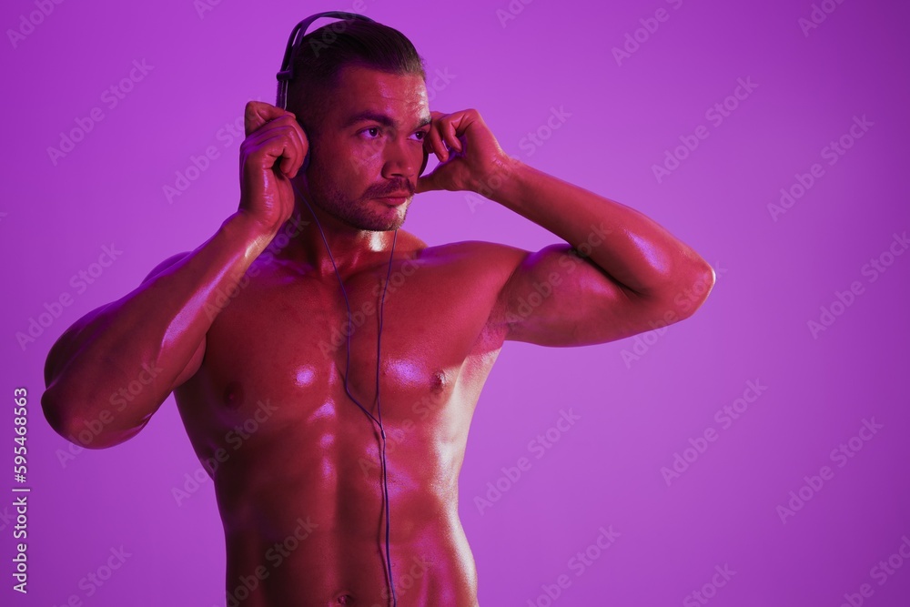 Fototapeta premium Man wearing headphones athletic physique bodybuilder nude torso abs full-length, fitness classes, purple colored light. Advertising, sports, active lifestyle, competition, challenge concept. 