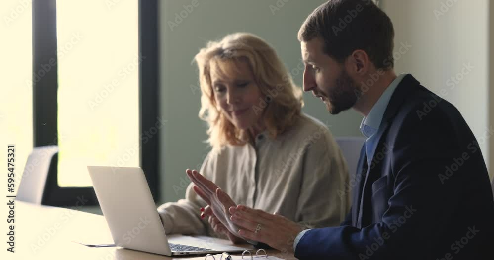 Serious confident business project leader explaining new employee work tasks, using laptop, pointing at display. Middle aged intern listening to speaking, mentor at workplace Serious confident business project leader explaining new employee work tasks, using laptop, pointing at display. Middle aged intern listening to speaking, mentor at workplace