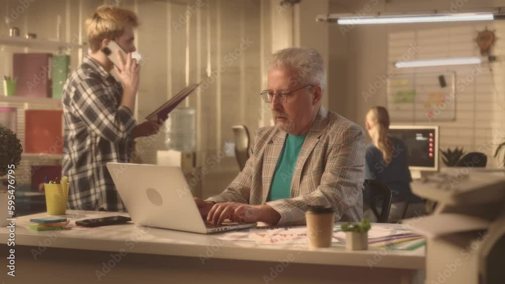 A man aged typing on a laptop keyboard. A businessman in a stylish jacket works in an office sitting at a table. A young creative team of managers are working in the background. A man aged typing on a laptop keyboard. A businessman in a stylish jacket works in an office sitting at a table. A young creative team of managers are working in the background.