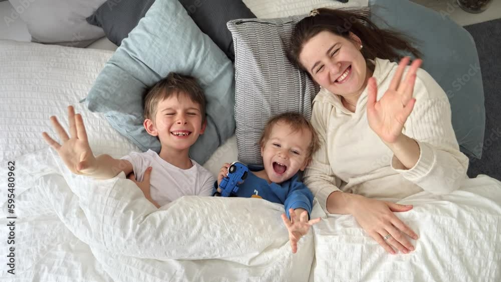 Young happy mother lying in bed with her two sons and waving in camera. Concept of family happiness, relaxing at home, having fun in bed, parent and cheerful kids Young happy mother lying in bed with her two sons and waving in camera. Concept of family happiness, relaxing at home, having fun in bed, parent and cheerful kids